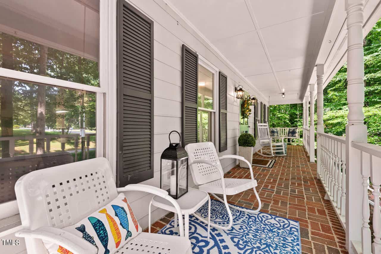 A cozy front porch with a white ceiling and brick flooring features white chairs, a blue patterned rug, and a lantern. There is a colorful fish-patterned pillow on one of the chairs. A green potted plant and lush greenery are visible in the background.