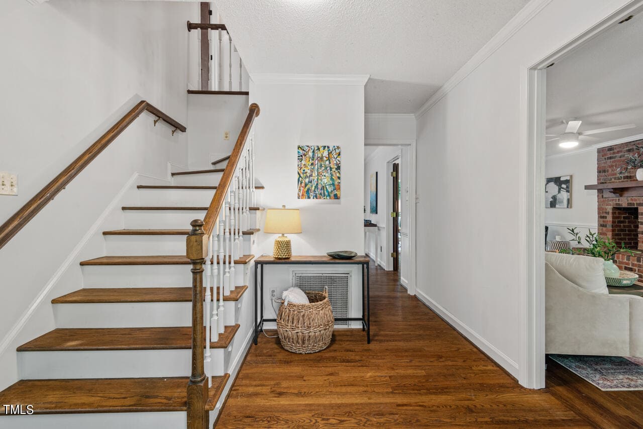A hallway with wooden floors leads to a staircase with wooden steps and white risers on the left. A console table with a lamp, decorative items, and a basket underneath stands against the wall. A doorway on the right reveals a living room with a brick fireplace.