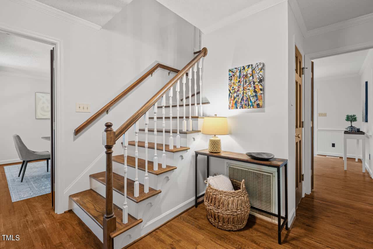 A modern home interior featuring a wooden staircase with white spindles leading to an upper floor. Below the stairs is a console table with a lamp, a decorative basket, and wall art. The space has wooden floors and white walls. A dining room is visible to the left.