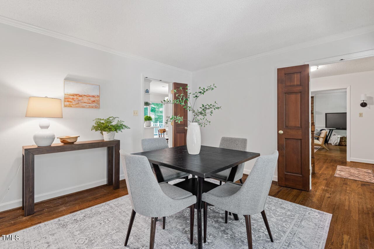 A modern dining room with a dark wooden table and four gray upholstered chairs on a patterned gray rug. The walls are white with one adorned by a framed piece of art. The room has a console table with decorative items, and a door opens to a living area.