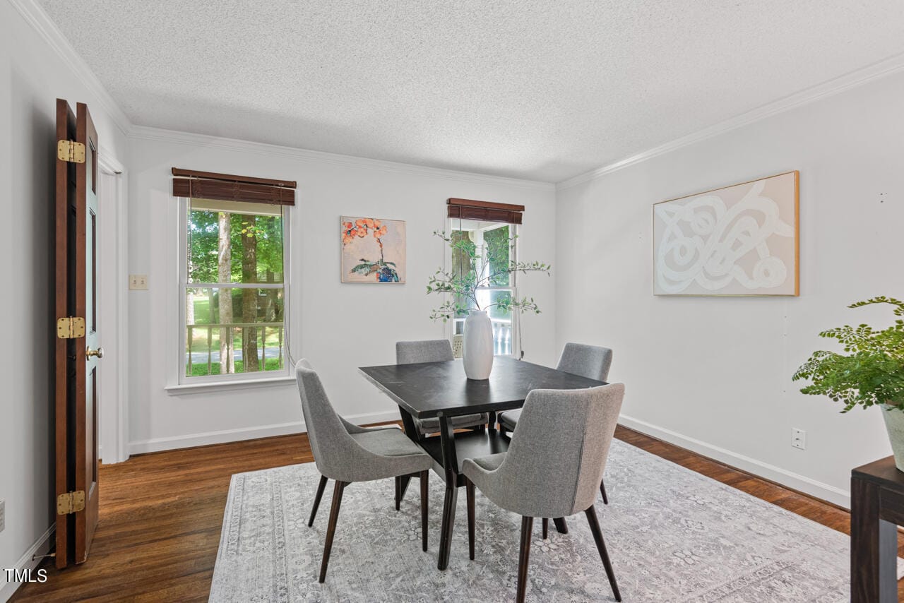 A modern dining room with hardwood floors, four gray upholstered chairs around a dark wood table, and white walls adorned with abstract art. Two windows with dark blinds let in natural light, and a potted plant sits in the corner on the right.