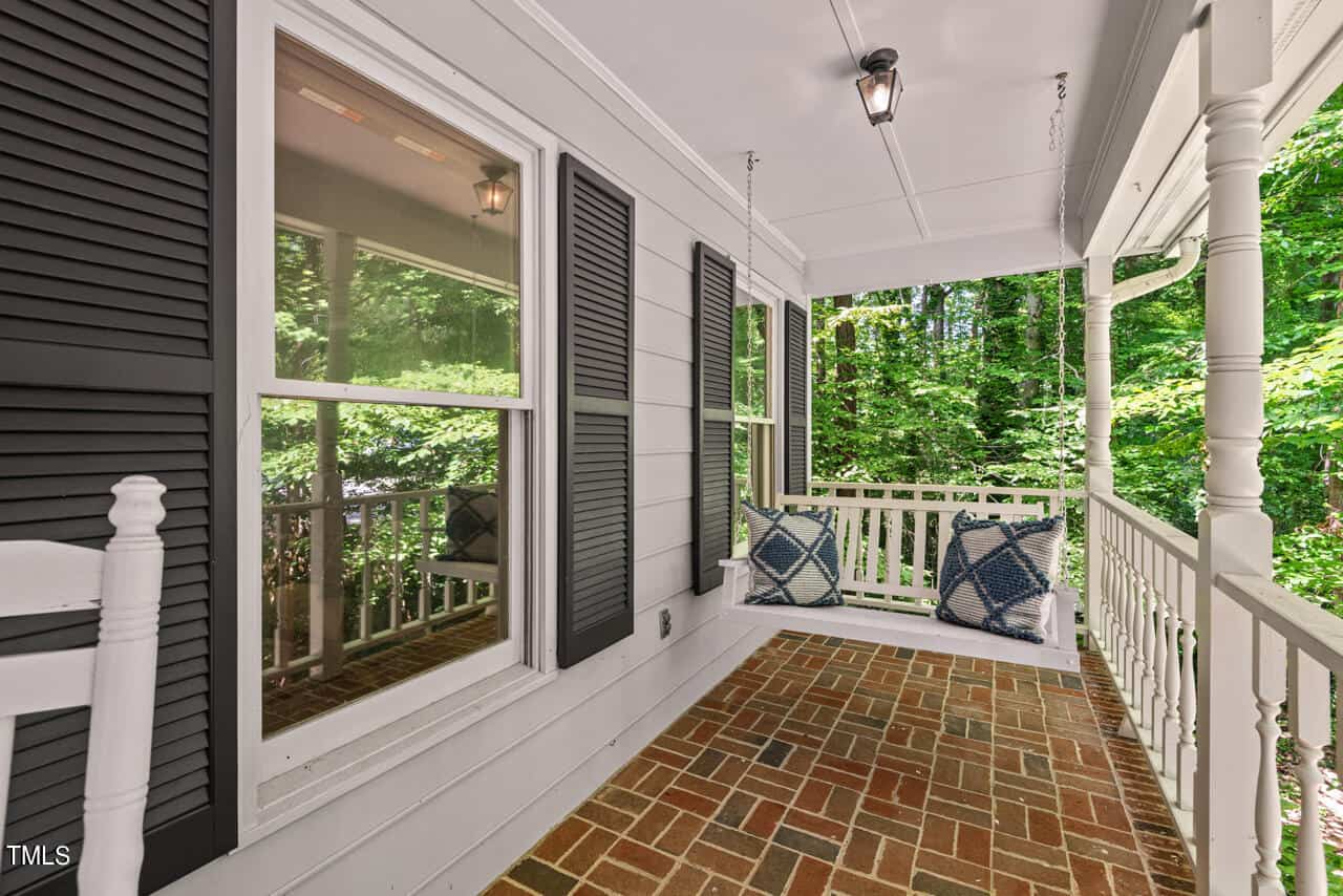 A cozy porch with a brick floor featuring a white porch swing adorned with two blue-and-white patterned pillows. The porch has white railing and black shutters on the windows, and is surrounded by lush greenery in the background.