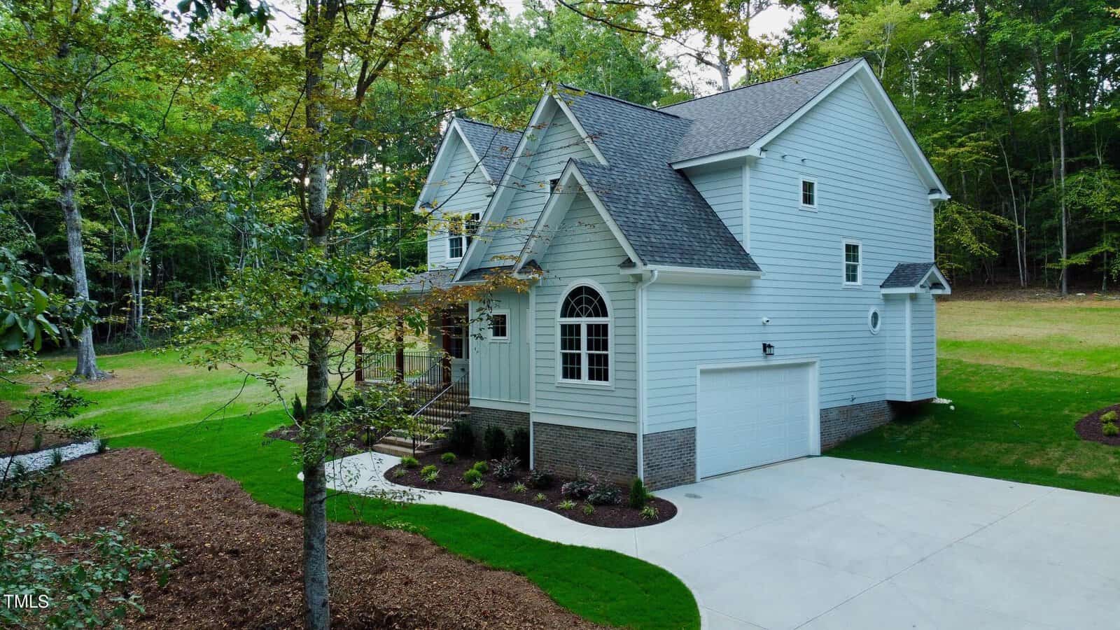 A two-story white house with a gabled roof, brick foundation, and attached two-car garage, surrounded by green lawn, trees, and landscaped garden beds. A concrete driveway leads up to the front entrance.
