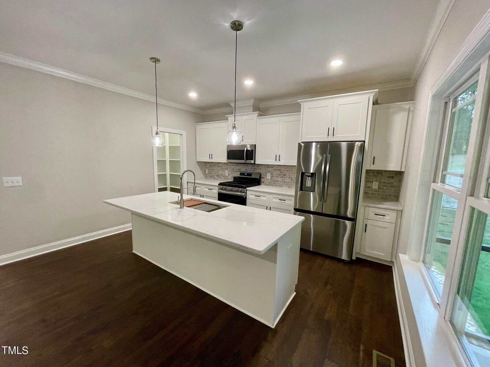 Modern kitchen with white cabinets, stainless steel appliances, a center island with sink and pendant lights, dark wood flooring, and large window providing natural light.