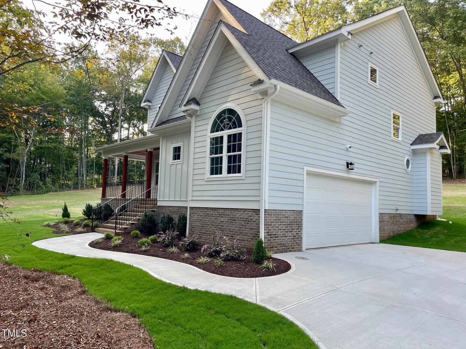 A modern light gray house with white trim, brick foundation, and attached two-car garage. A curved concrete walkway and landscaped garden beds border the green lawn. Trees are visible in the background.