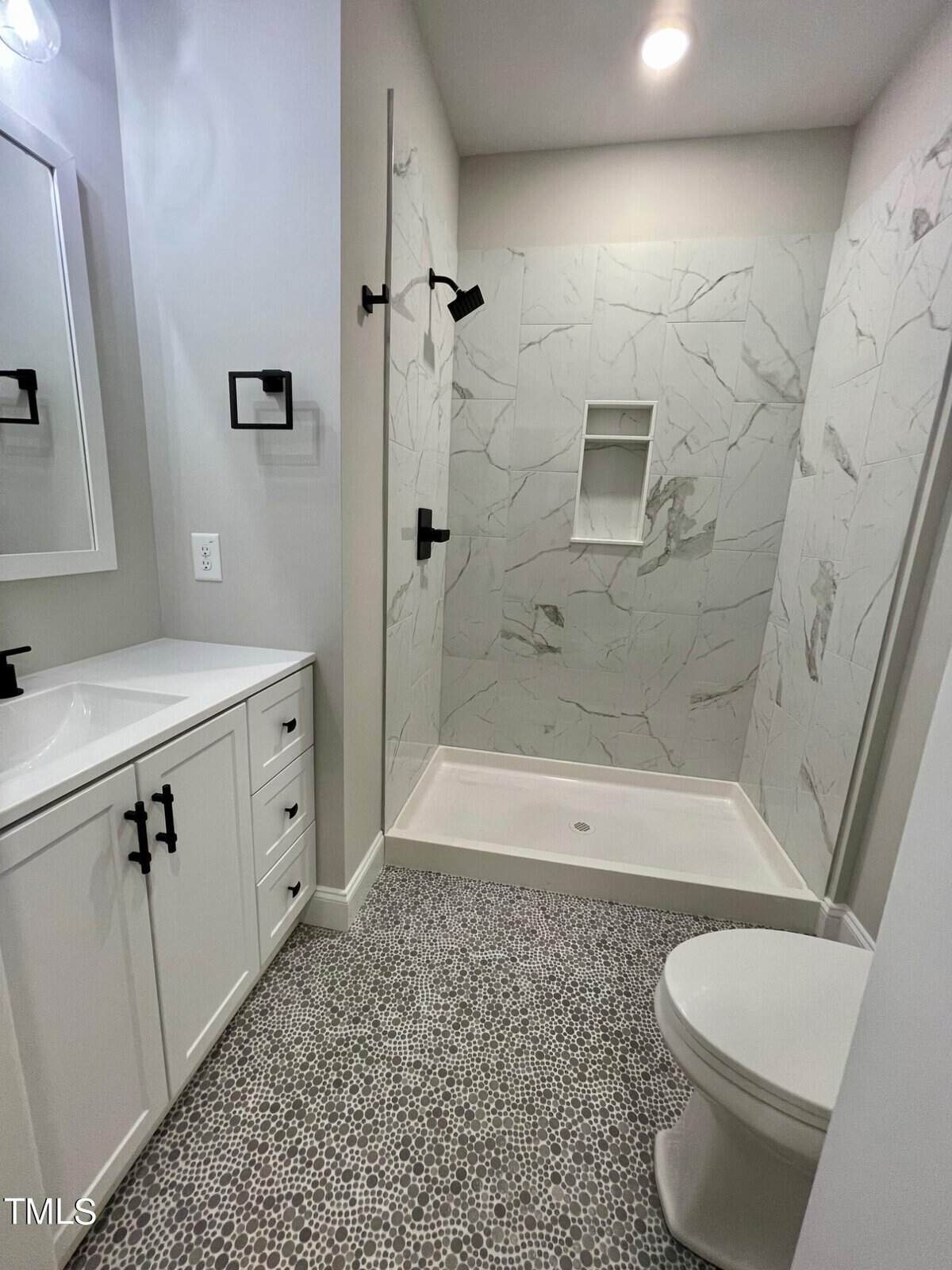 Modern bathroom with a walk-in shower featuring white marble-patterned walls, built-in shelf, and black fixtures. A white vanity with black handles and a round toilet sit on a gray pebble-patterned tile floor.