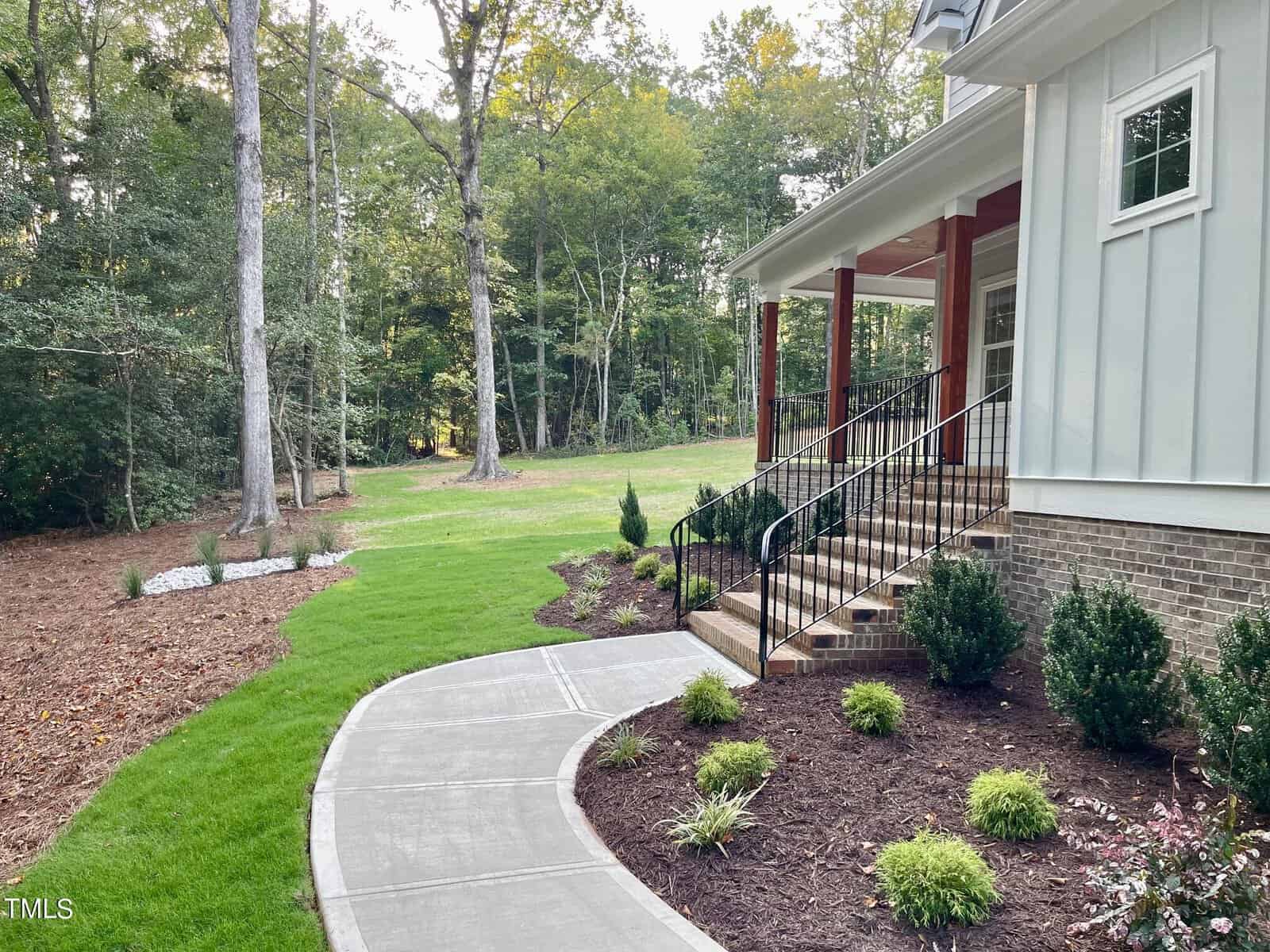 A concrete walkway curves past trimmed bushes and a landscaped yard leading to the steps of a house with a covered porch, surrounded by tall trees and a grassy lawn.