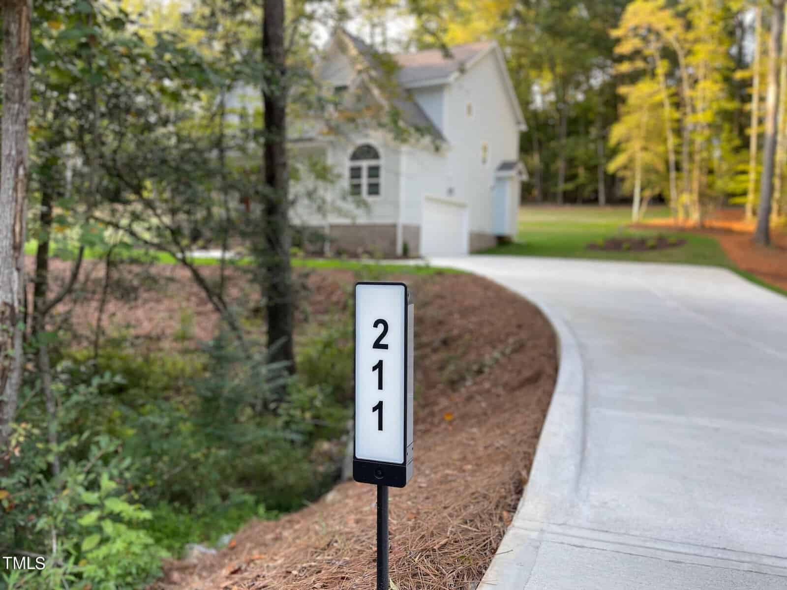 A driveway curves toward a white house surrounded by tall trees. In the foreground, a sign with the number 211 is mounted next to the driveway.