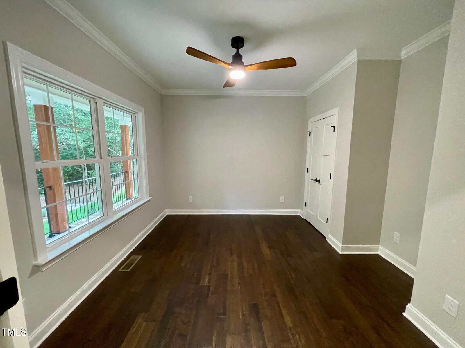 A small empty room with light gray walls, dark wood floors, a ceiling fan, a large window on the left letting in natural light, and double white closet doors.