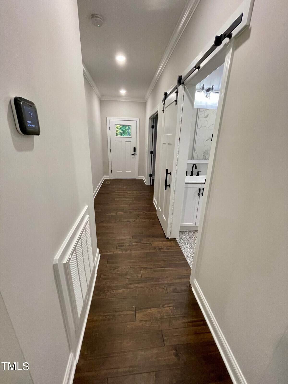A hallway with dark wood floors, white walls, and recessed lighting. A sliding barn door reveals a bathroom with a white vanity. At the end of the hall is a white front door with a window. A thermostat is on the left wall.