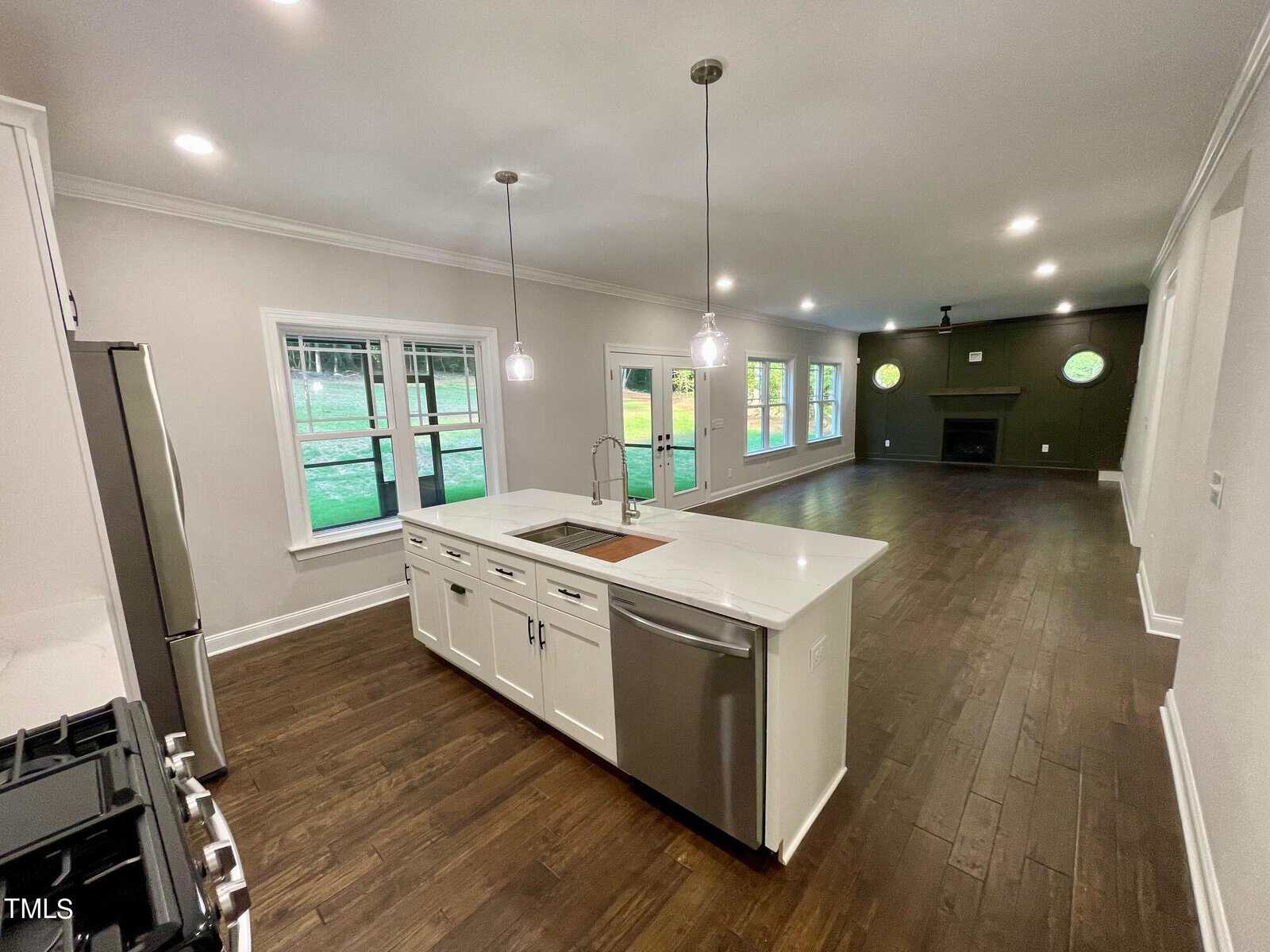 Modern open-concept kitchen and living room with dark hardwood floors, a large island with a sink, stainless appliances, pendant lights, and windows overlooking a green lawn.
