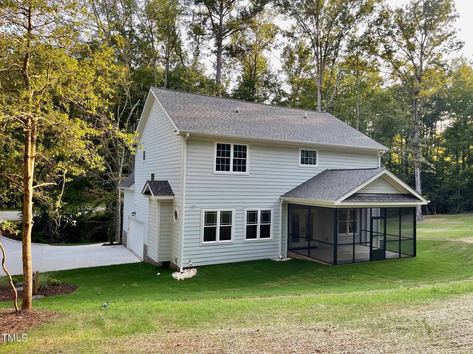 Two-story white house with gray roof, attached garage, and a screened-in back porch. The house sits on a grassy lawn and is surrounded by tall trees and a wooded area.