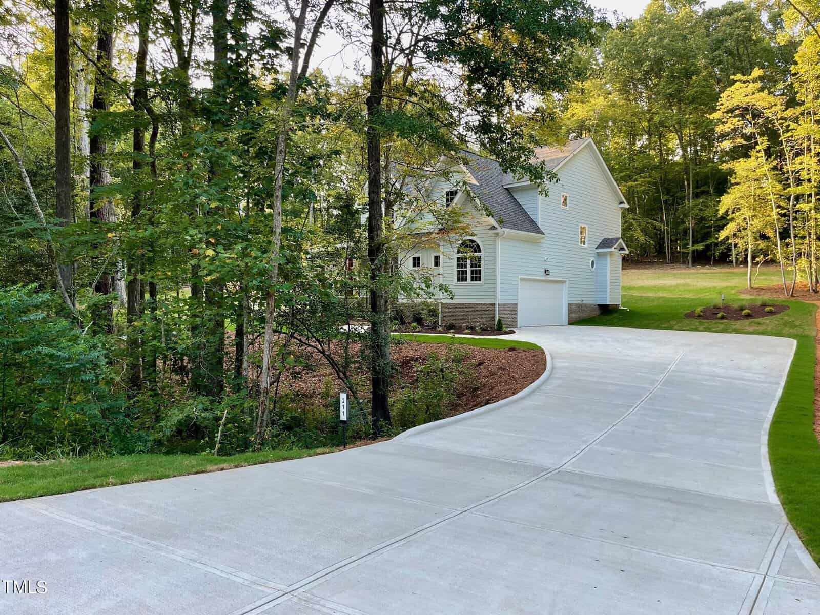 A white two-story house with a gray roof sits near a wooded area, at the end of a long, curved concrete driveway. Trees and landscaped grass surround the property under a clear sky.