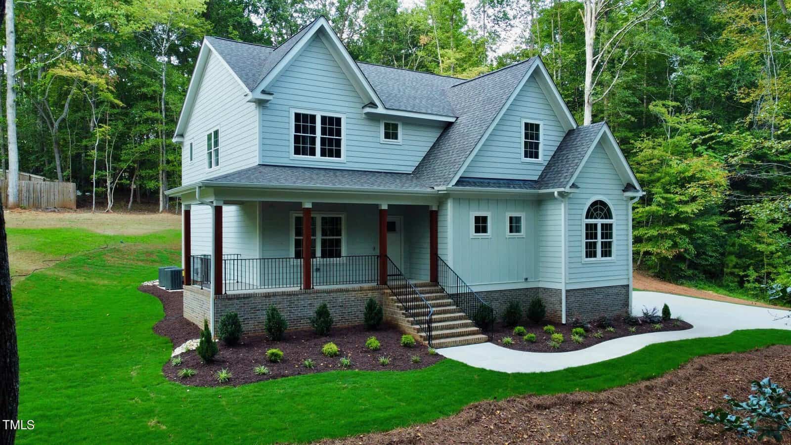 Two-story light gray house with a covered front porch, dark roof, and red front door, surrounded by freshly landscaped yard, green grass, and tall trees in the background. Concrete driveway curves along the right side.