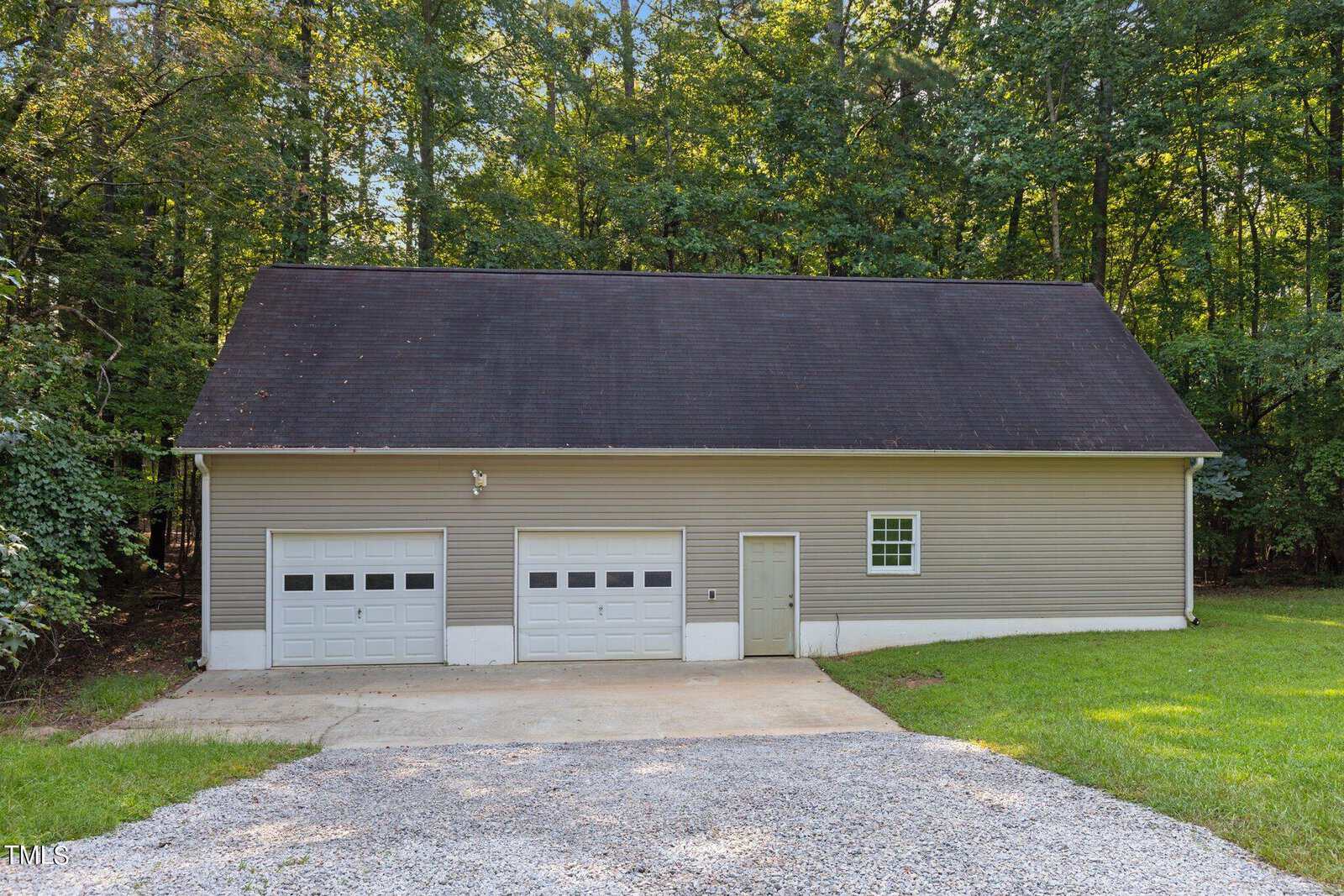 A beige detached garage with two white garage doors, a single entry door, and one window sits on a concrete driveway with a gravel parking area, surrounded by green grass and trees in the background.