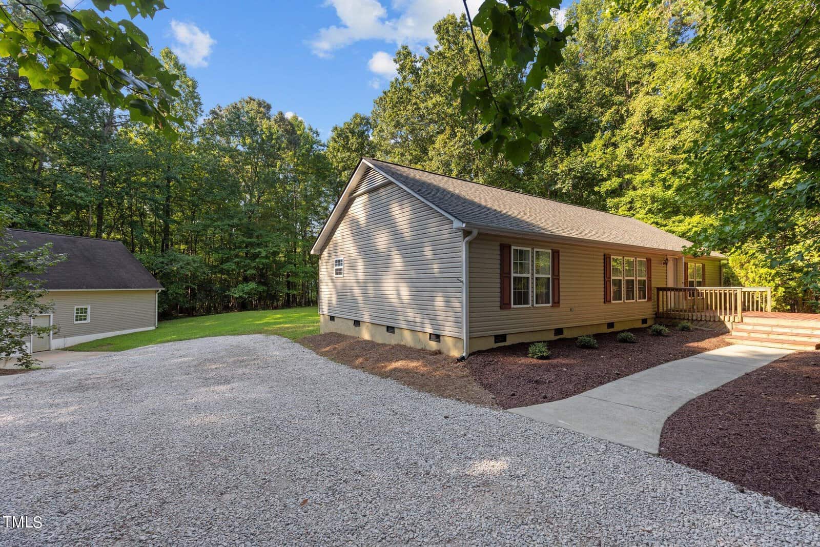 A single-story beige house with white trim sits next to a gravel driveway and a walkway. Trees surround the property, and a detached garage is visible in the background. The sky is mostly clear.