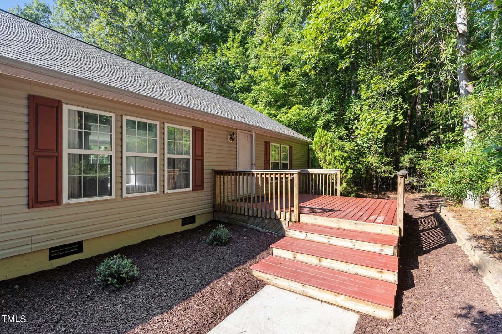 A beige, single-story house with red shutters is shown. A wooden porch with steps leads to the front door. The yard has mulch, small bushes, and is bordered by dense green trees.