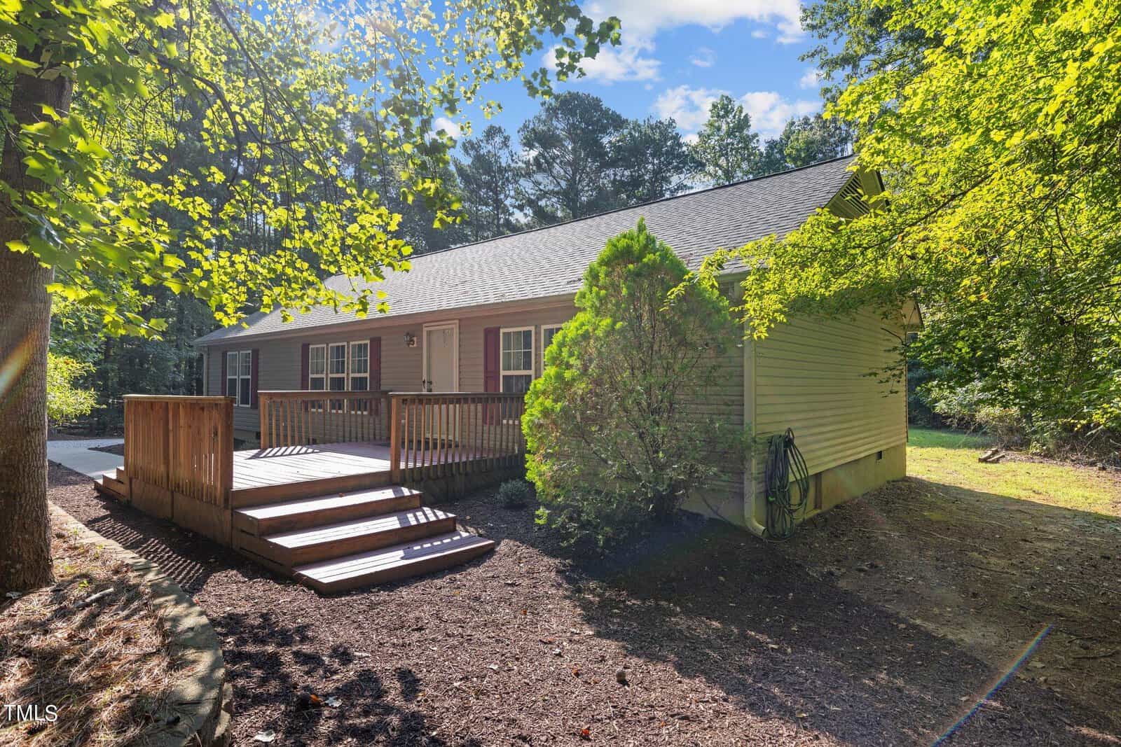 Single-story house with beige siding, a dark gray roof, and a wooden ramp leading to the front entrance. The house is surrounded by trees and bushes, with sunlight filtering through the branches.