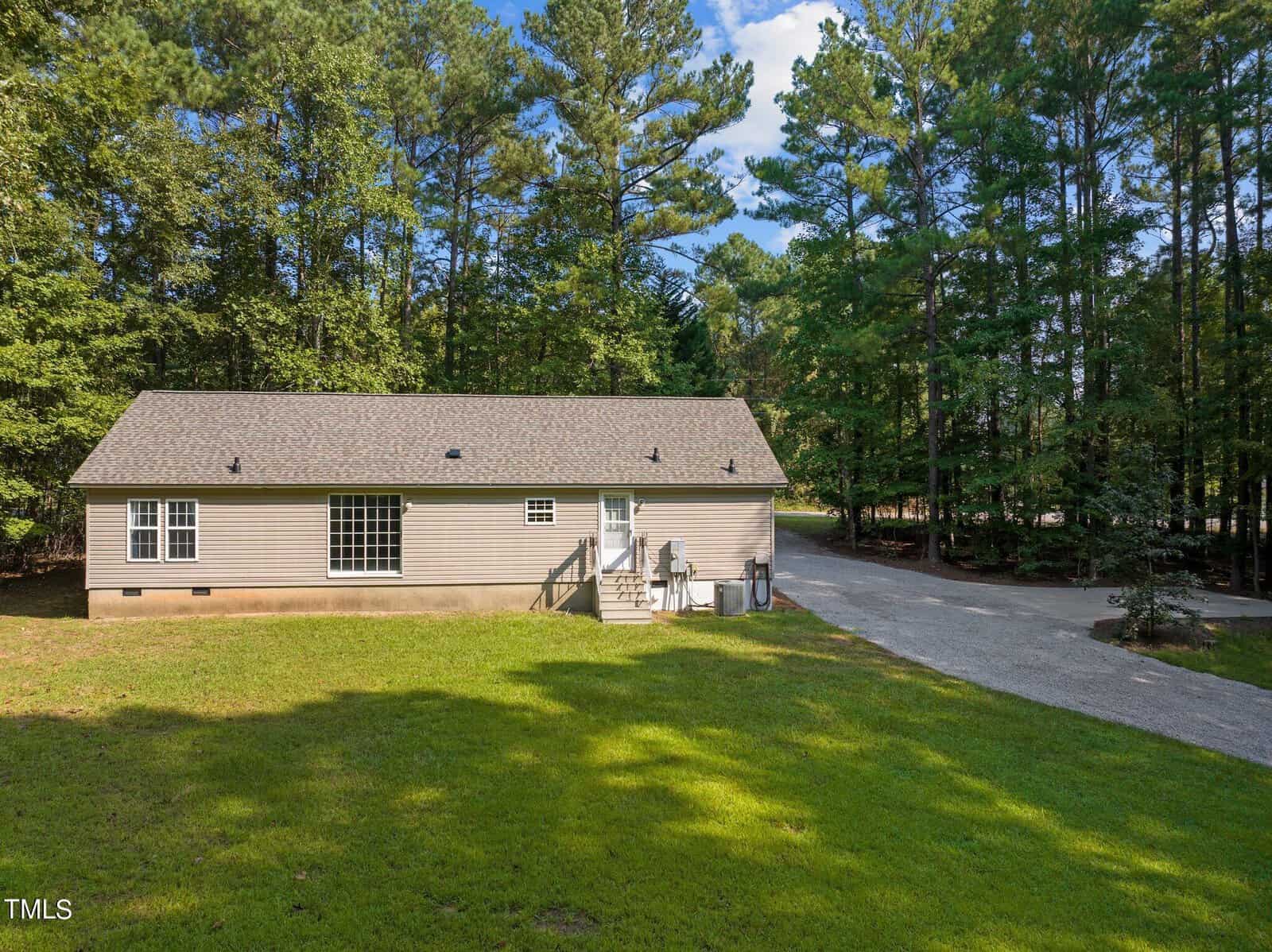 A single-story beige house with a shingle roof sits on a grassy lot bordered by tall trees. A few steps lead to the back door, and an air conditioning unit is visible near a gravel driveway.