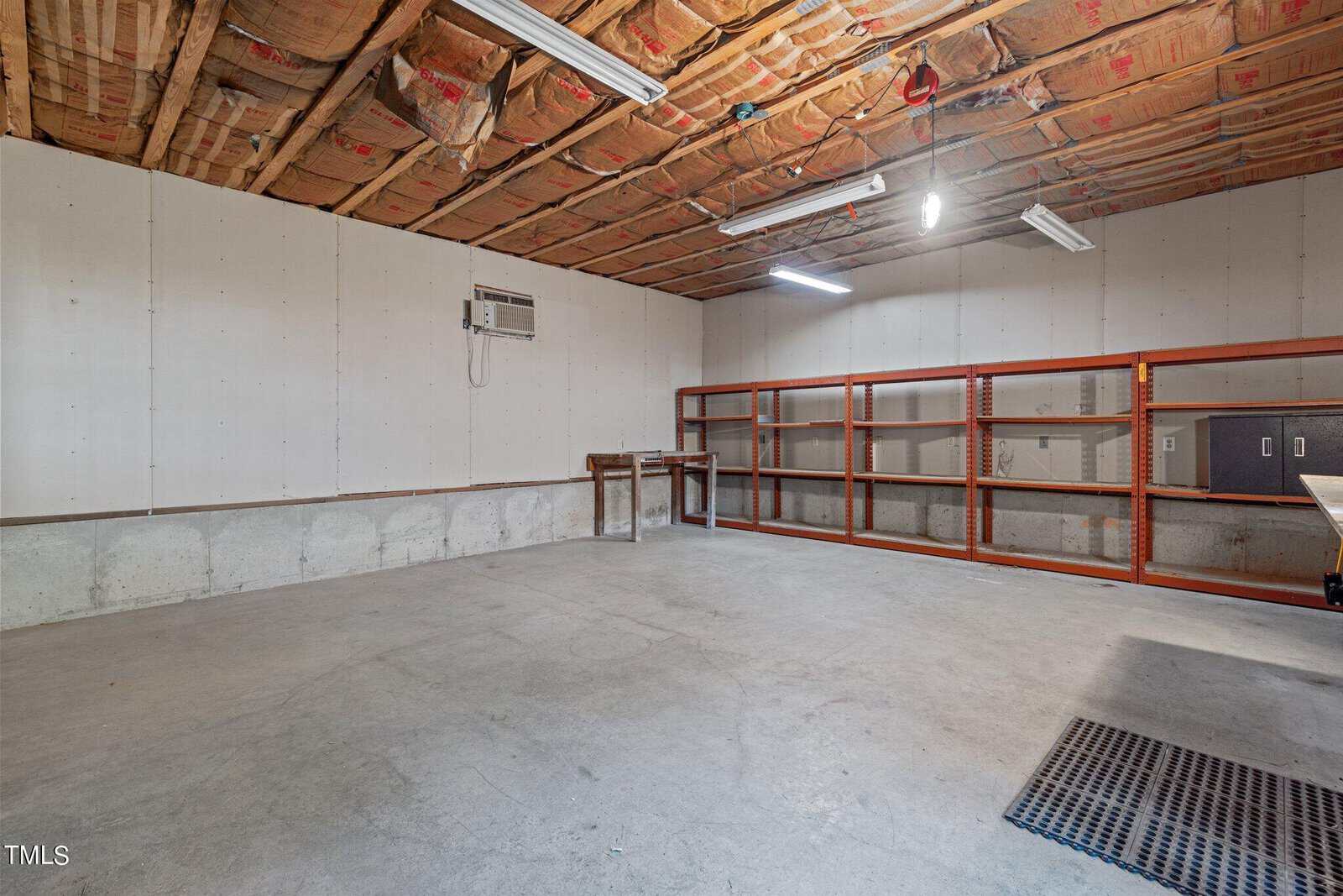 Spacious unfinished basement with concrete floor, exposed insulation on the ceiling, wall-mounted air conditioner, fluorescent lights, and metal shelves along one wall.