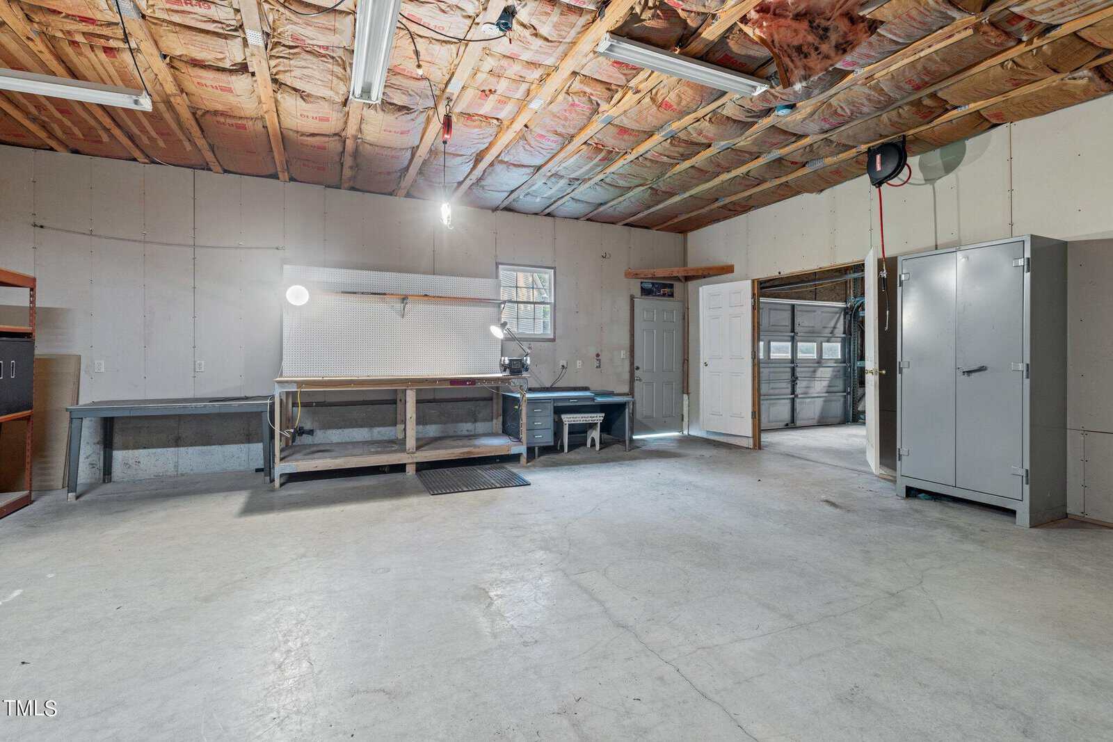 A spacious garage with exposed insulation on the ceiling, concrete floors, workbenches along the walls, a metal storage cabinet, a small window, overhead lights, and an open garage door in the background.