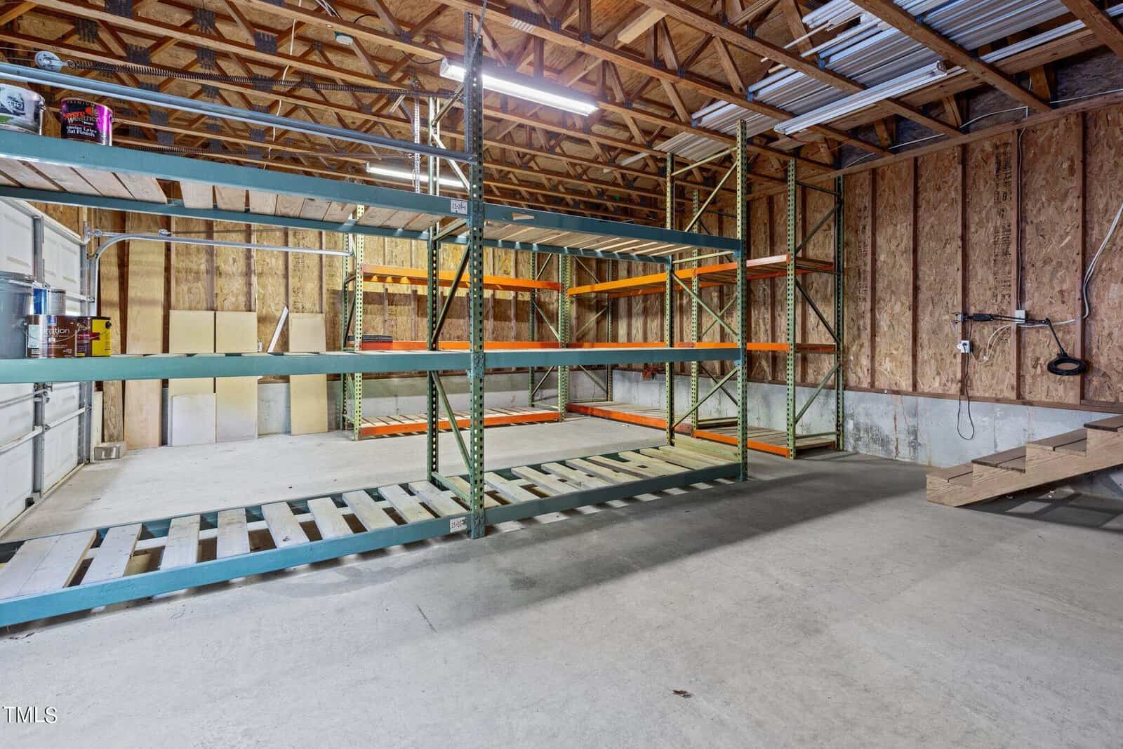 Large garage or warehouse interior with exposed wooden beams, metal shelving racks, unfinished walls, a concrete floor, and a staircase leading up to a loft or second level on the right.