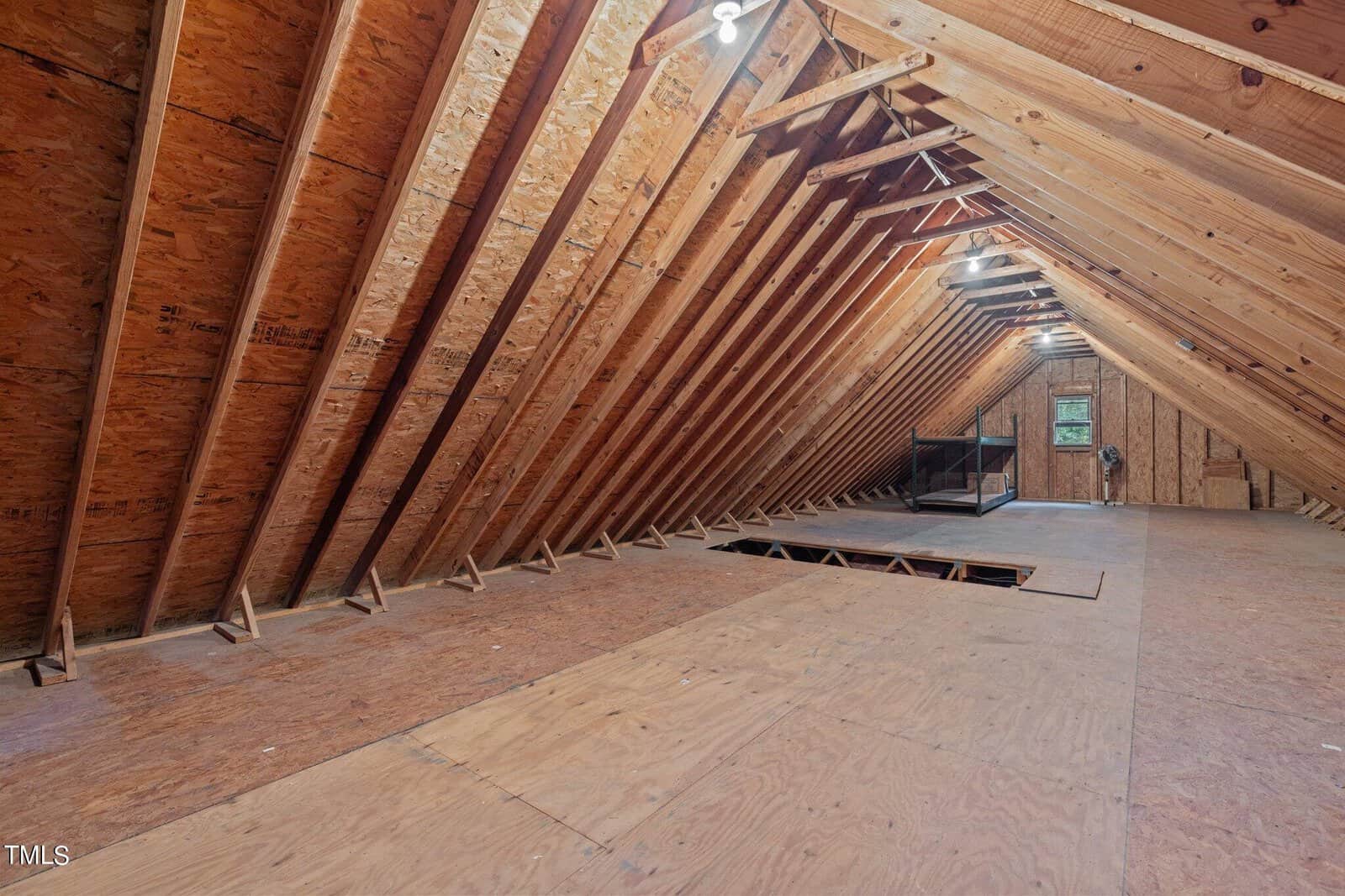 Unfinished attic with exposed wooden beams, plywood flooring, and a rectangular hole in the floor. There is a window and some furniture in the back. Ceiling lights are turned on, illuminating the space.