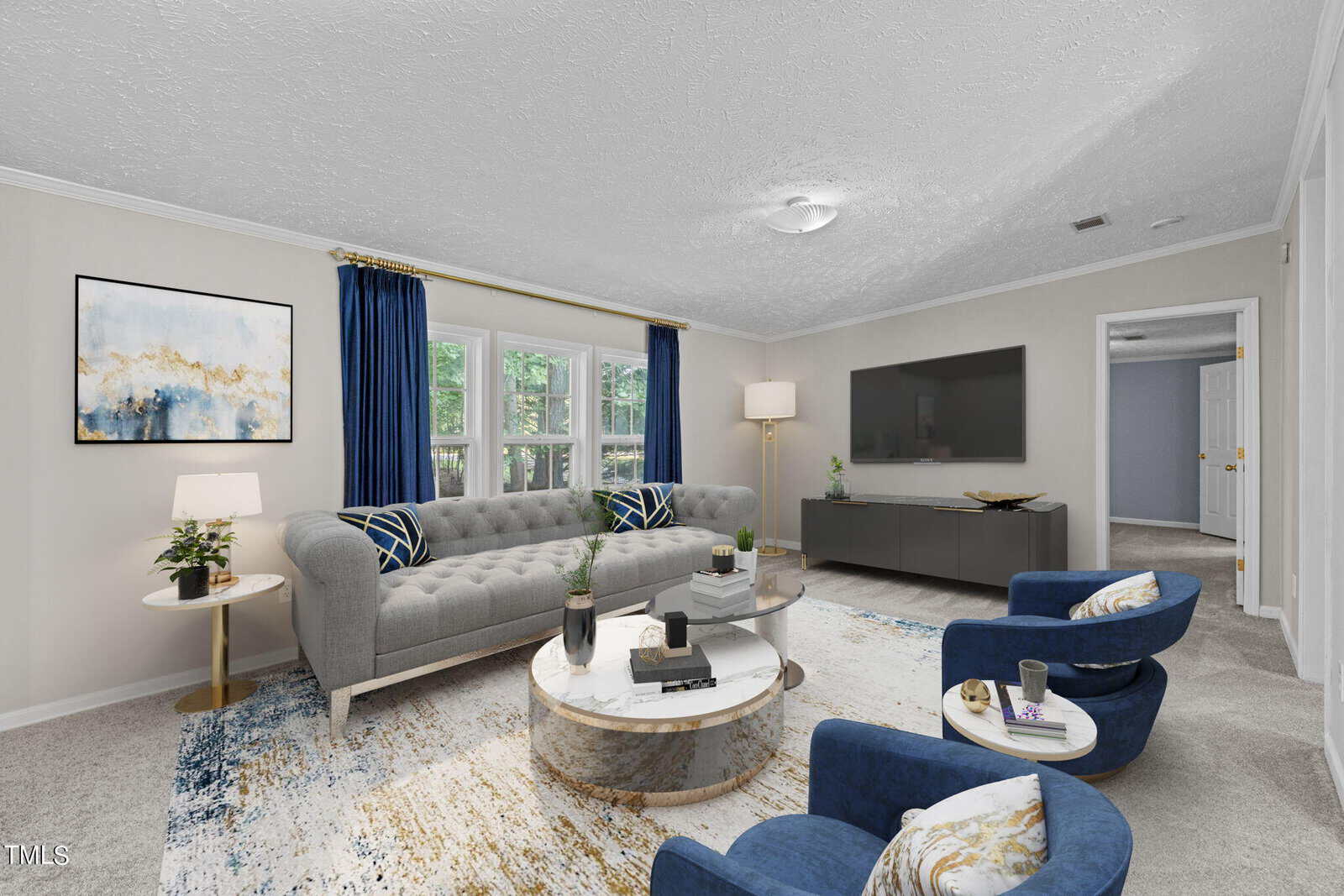 A living room with a gray tufted sofa, two blue chairs, a round coffee table, a TV on a console, large windows with blue curtains, potted plants, and abstract wall art on light-colored walls.