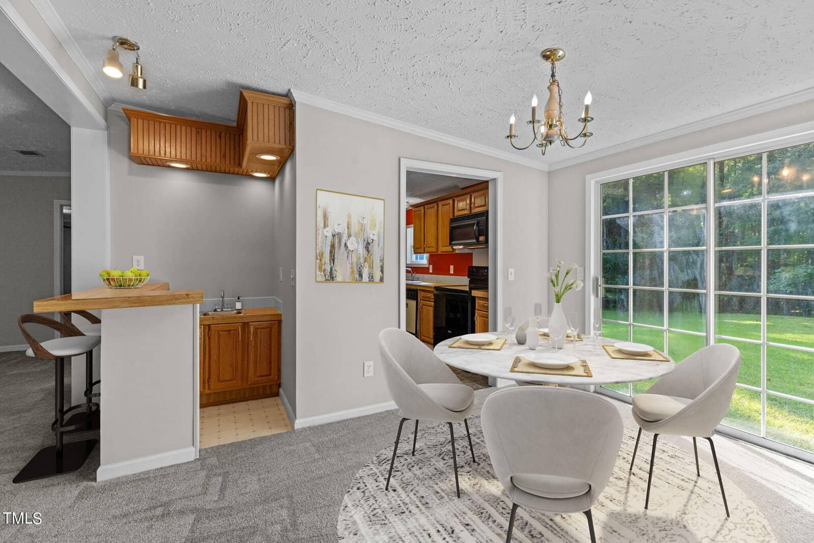 A dining area with a round table set for four, white chairs, a chandelier, and large windows overlooking a green lawn. A wet bar with wooden cabinets and a small kitchen are visible in the background.