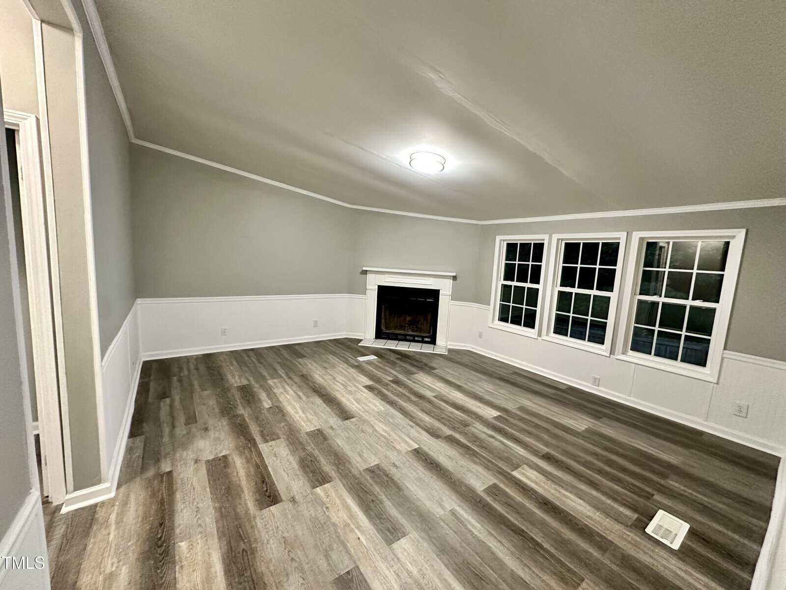 A spacious living room with light gray walls and wood-paneled flooring. There is a white fireplace against the far wall, beneath a ceiling light. Three windows are on the right, letting in natural light. A white chair rail runs around the room.
