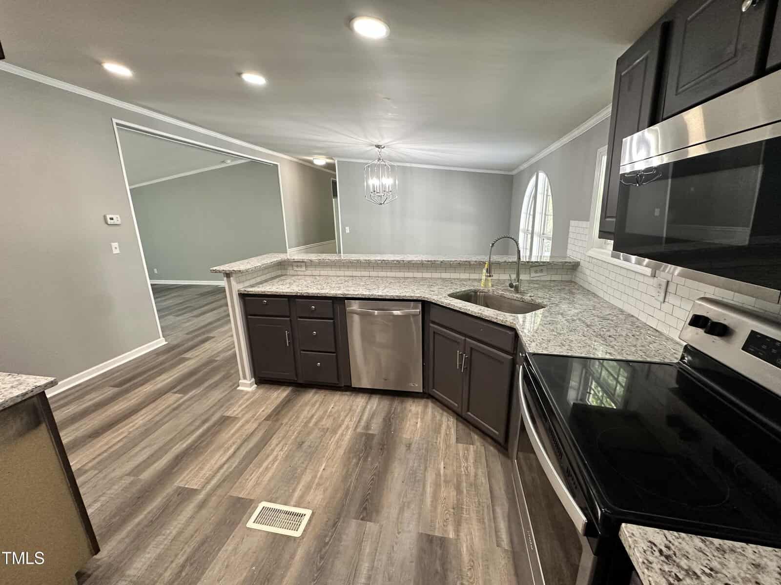 Modern kitchen with dark cabinets, granite countertops, and stainless steel appliances. The open layout leads to a well-lit living area with a chandelier. Light wood flooring and a window add to the ambiance.