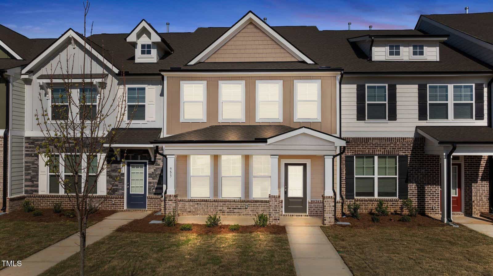 A two-story townhouse with beige siding, brick accents, white trim, and black shutters. The front door and windows face a small, landscaped yard with a sidewalk leading to the entrance.
