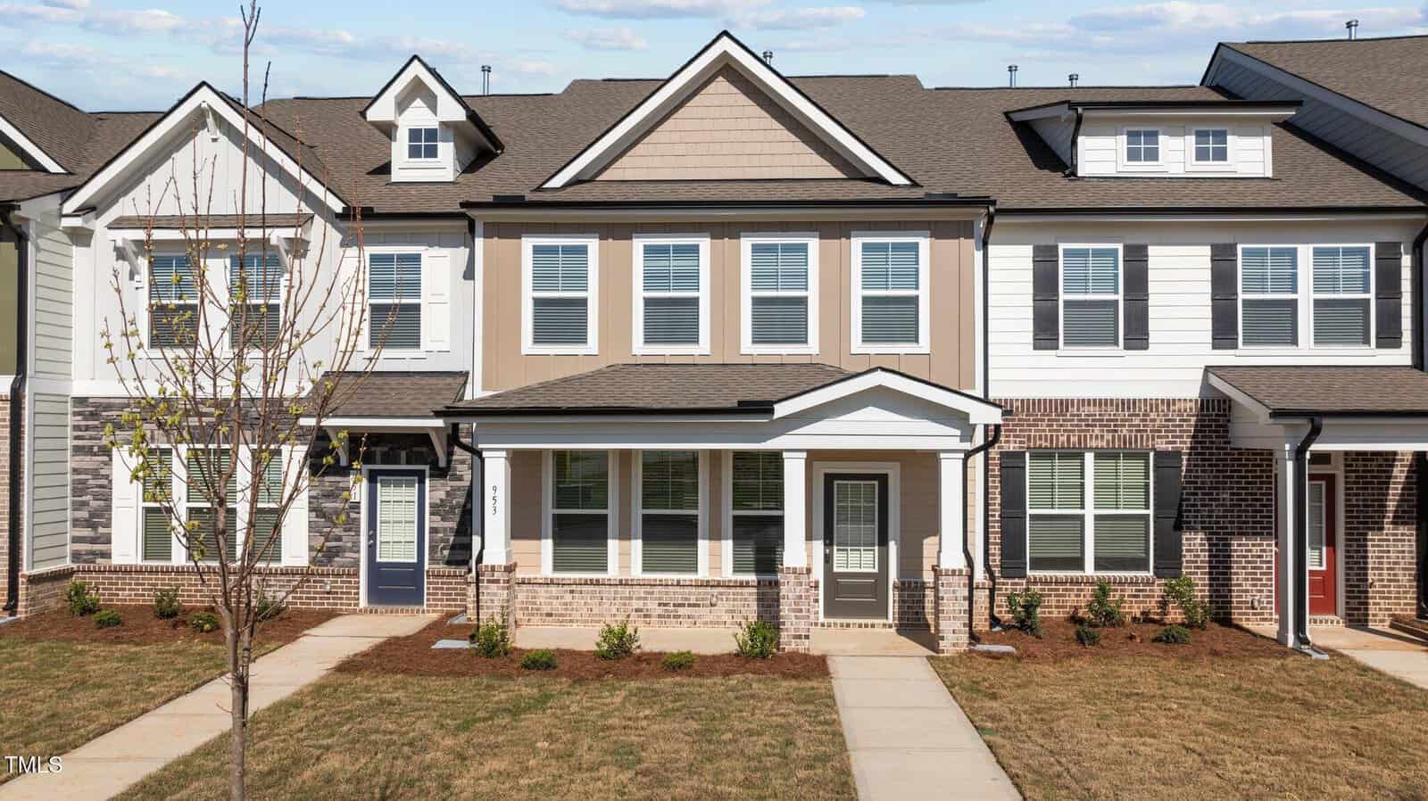A row of modern two-story townhomes with beige siding, white trim, and brick accents. The middle unit has a covered entryway, multiple windows, and a small front yard with minimal landscaping.