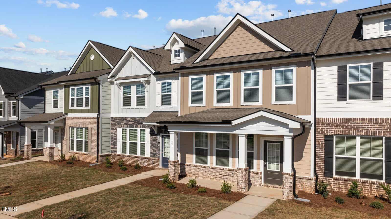 A row of modern two-story townhomes with varied siding colors and brick accents, featuring white-trimmed windows, front doors, and small landscaped yards under a blue sky with scattered clouds.