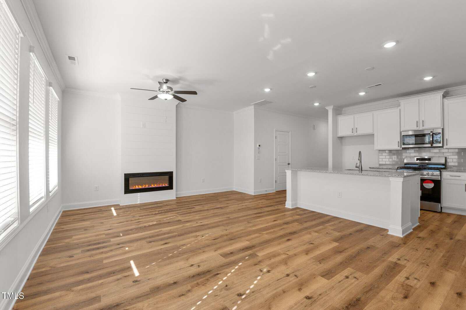 Unfurnished open-concept living area with wood flooring, white walls, a ceiling fan, a built-in wall fireplace, large windows, and a kitchen with white cabinets, stainless steel appliances, and an island.