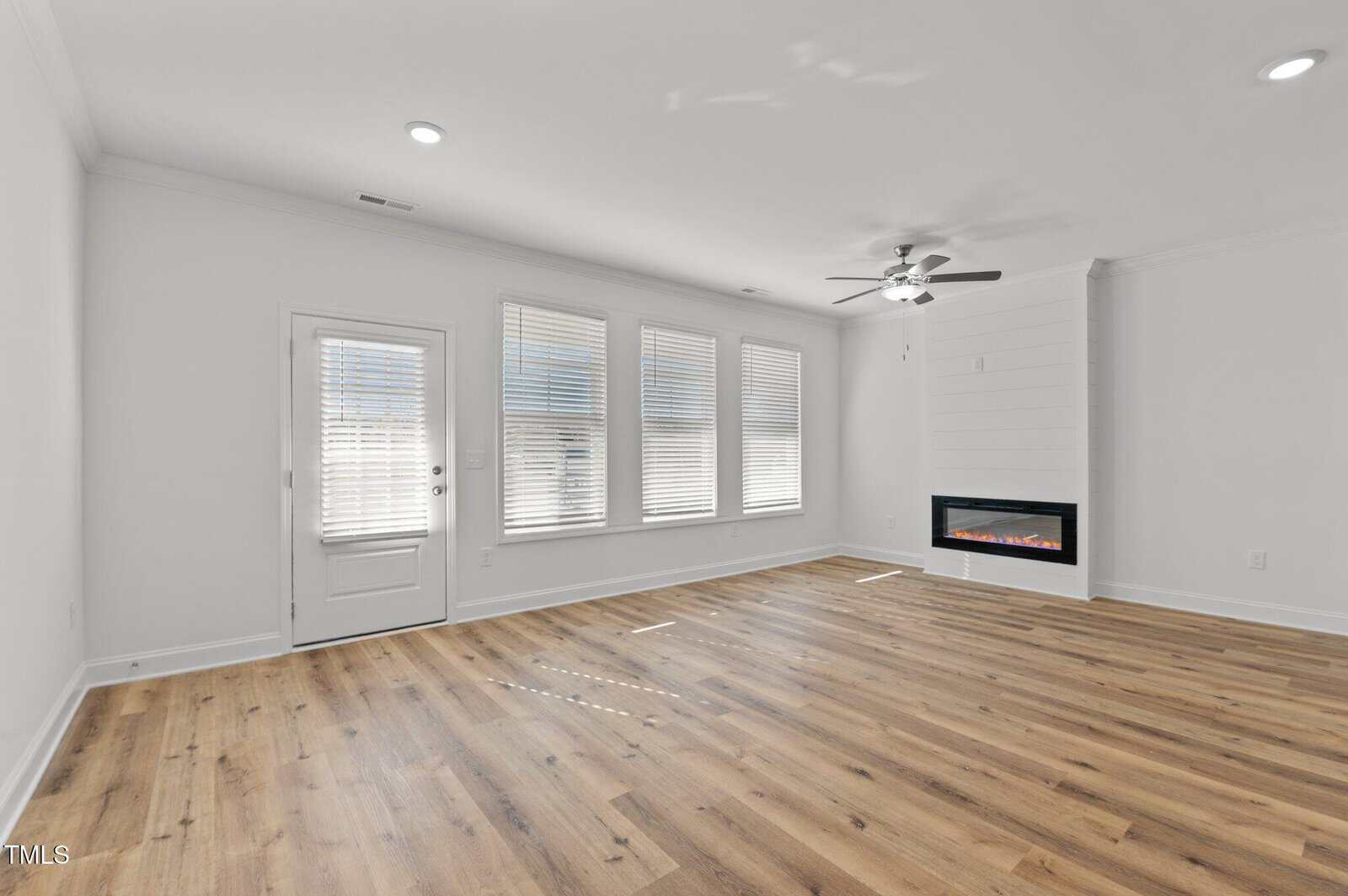 A bright, empty living room with light-colored walls, three large windows with blinds, a ceiling fan, a modern wall-mounted fireplace, and wood-style flooring. A door to the left opens outside.