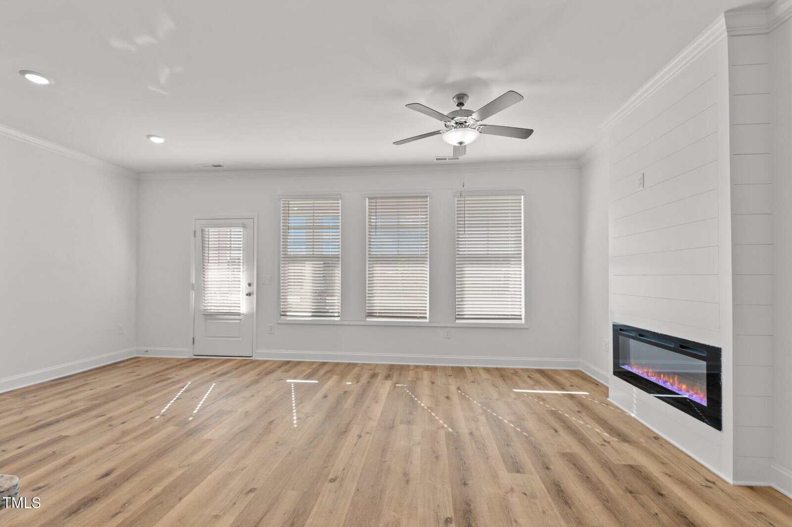 A bright, empty living room with light wood flooring, white walls, three large windows with blinds, a ceiling fan with light, a door, recessed lighting, and a modern electric fireplace.