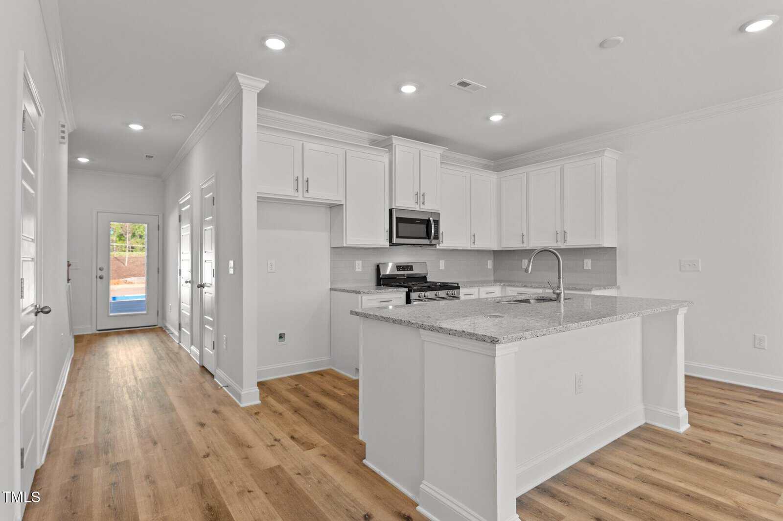 A modern kitchen with white cabinets, stainless steel appliances, a built-in microwave, a gas stove, a granite island with a sink, and light wood floors; hallway and door visible in the background.