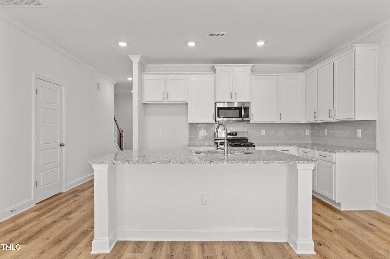 A modern kitchen with white cabinets, granite countertops, a center island with a sink, stainless steel appliances including a microwave and oven, subway tile backsplash, and light wood flooring.
