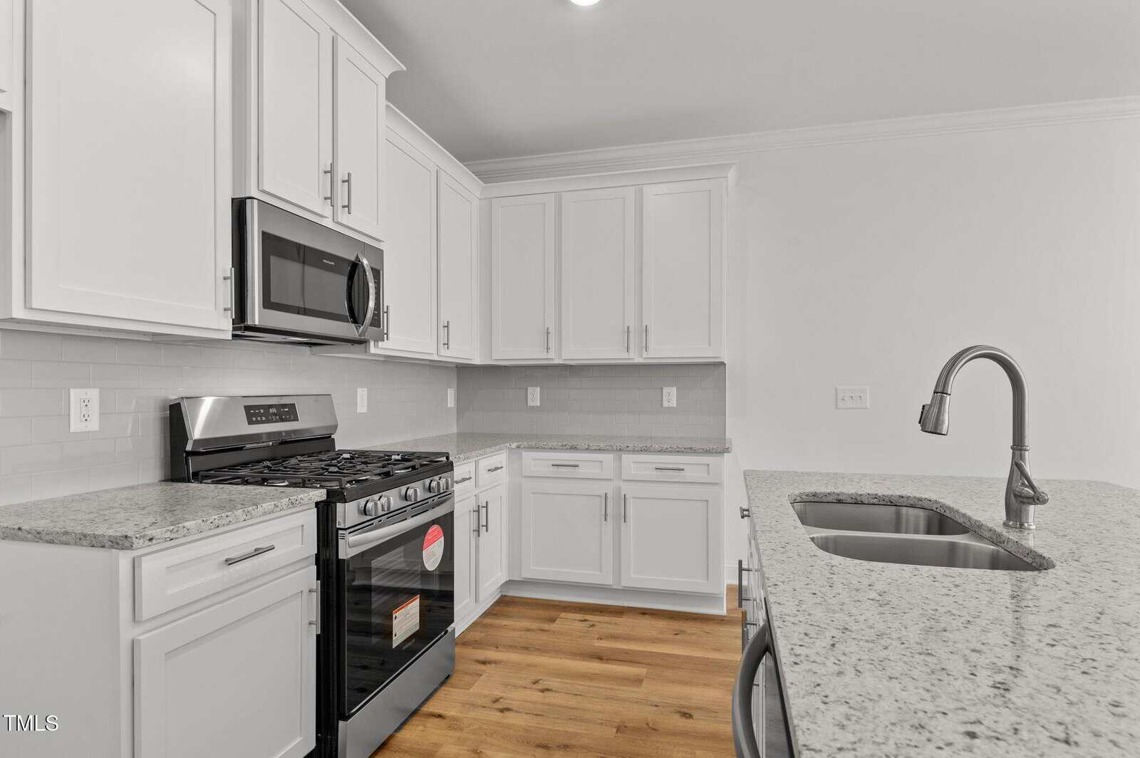 A modern kitchen with white cabinets, granite countertops, a stainless steel gas stove and oven, a built-in microwave, a double-basin sink with a faucet, and light wood flooring.