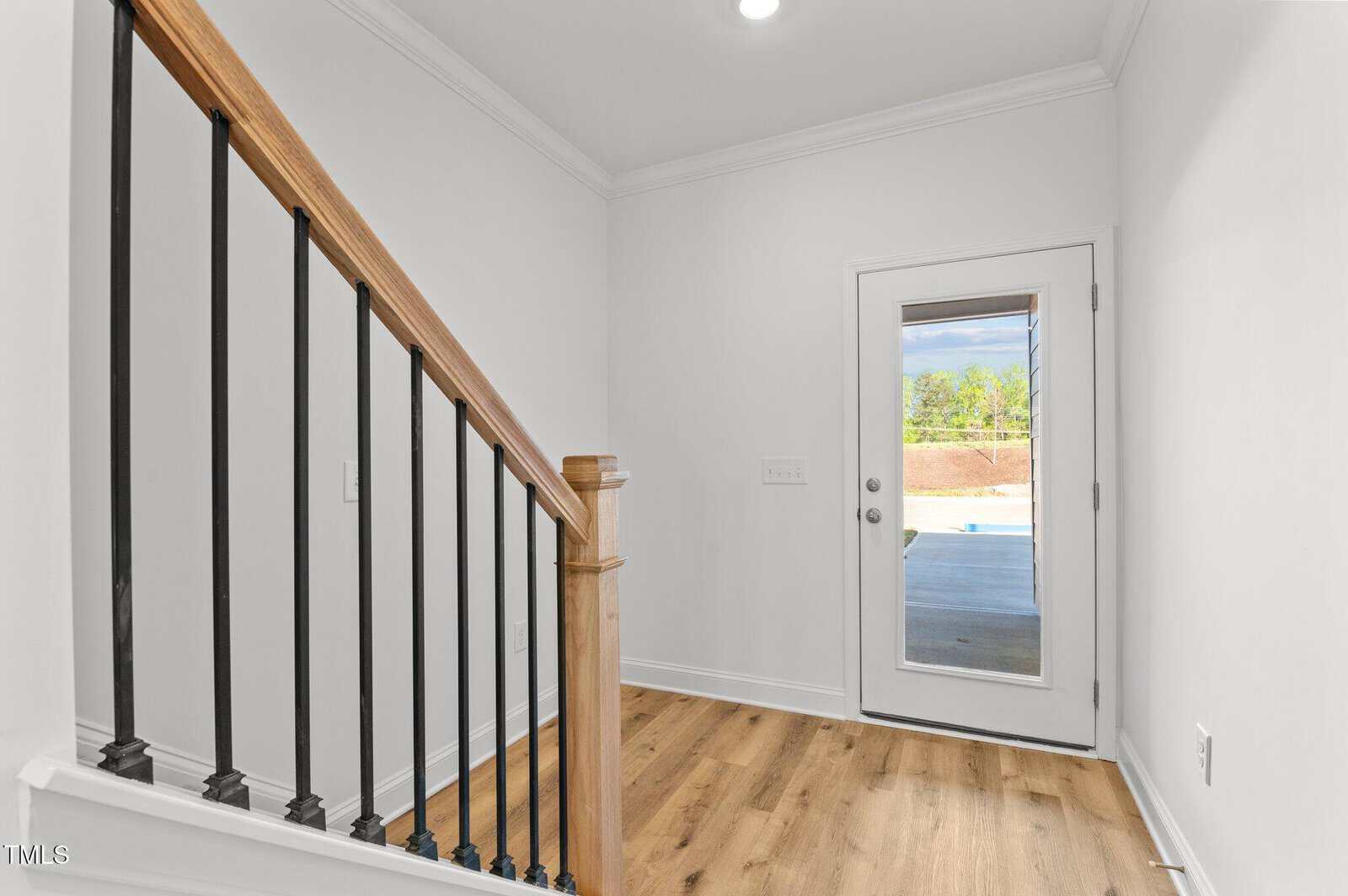 A small entryway with light wood flooring, a wooden staircase with black metal railings on the left, white walls, and a glass door leading outside to a backyard with a covered patio and visible greenery.