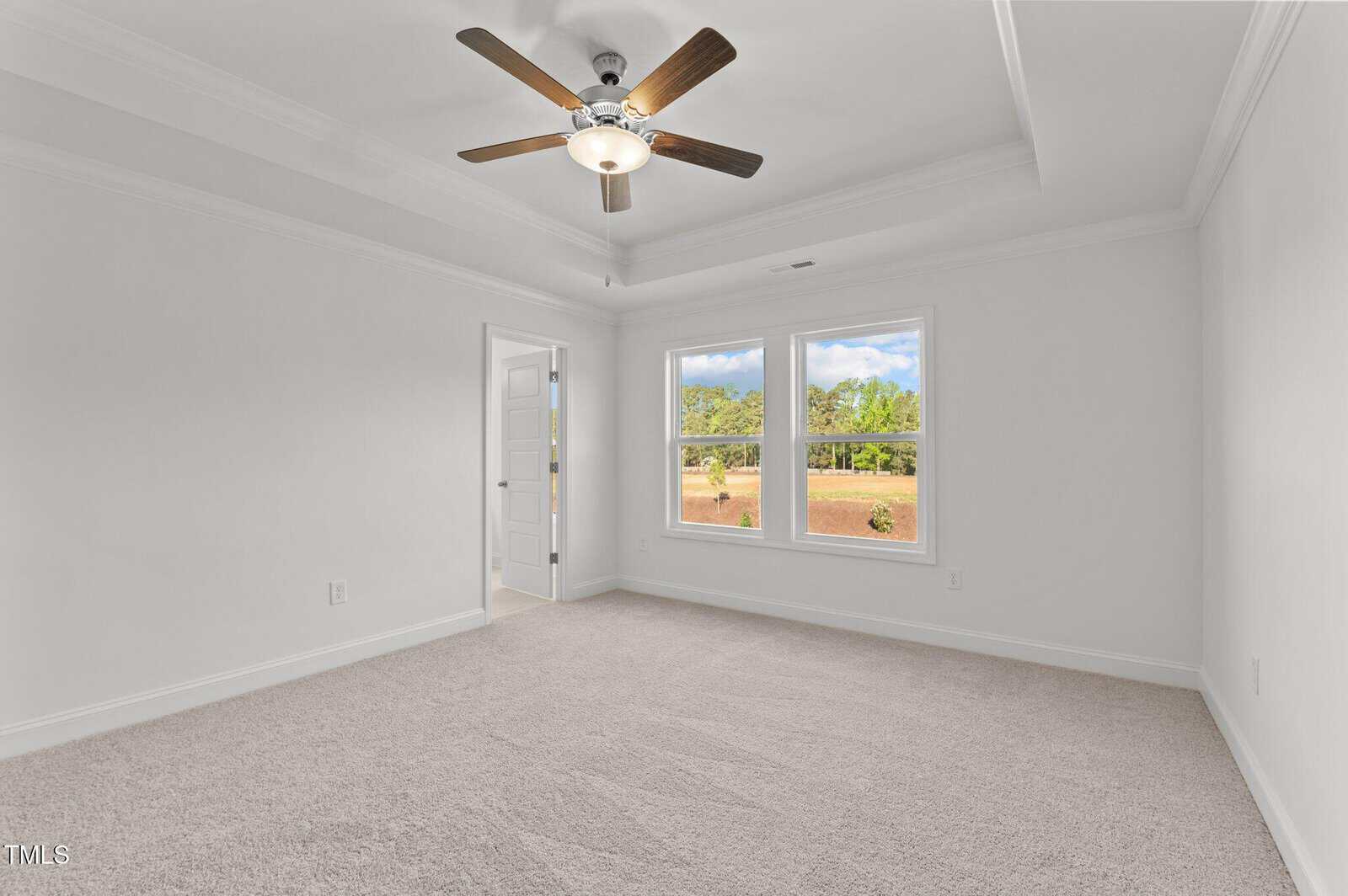 Bright, empty bedroom with beige carpet, white walls, ceiling fan, two large windows showing greenery, and a door leading to another room.