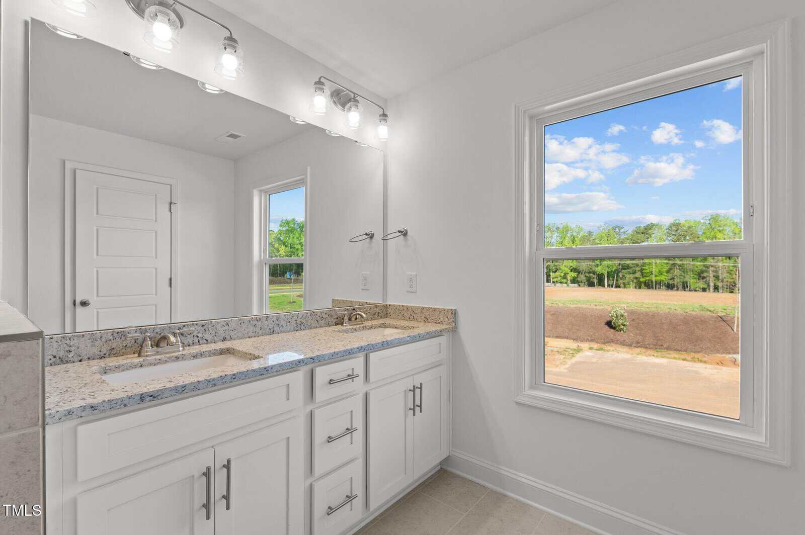Bright bathroom with white cabinets, granite countertop, double sinks, large mirror with overhead lights, and a window showing a clear sky and greenery outside.