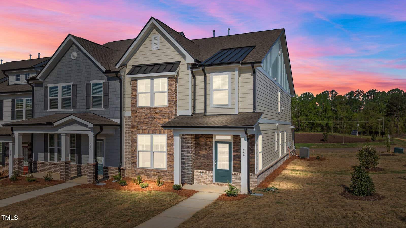 A two-story townhouse with gray siding, brick accents, white trim, and black roof, sits on a corner lot with a pathway leading to the front door. The sky is pink and blue at sunset, and the yard is mostly grass with minimal landscaping.