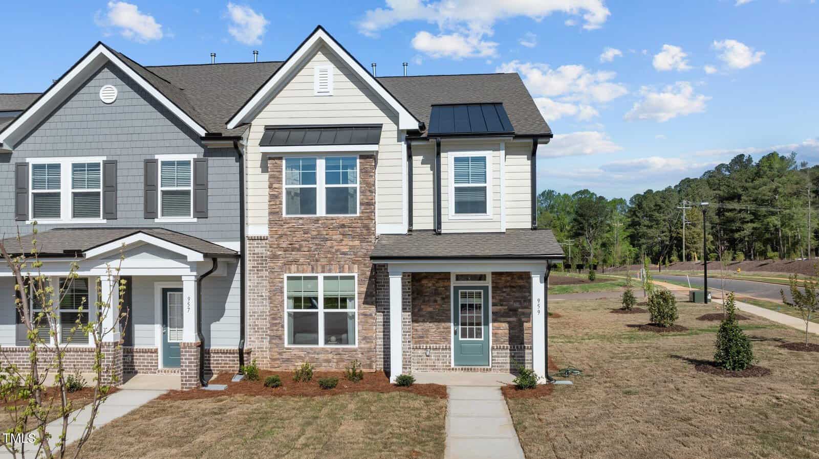 Two-story townhouse with gray siding, brick accents, and white trim. Front door has a glass panel. Adjacent unit is attached. The lawn is sparse with young trees; sidewalks run along the front and side of the property.
