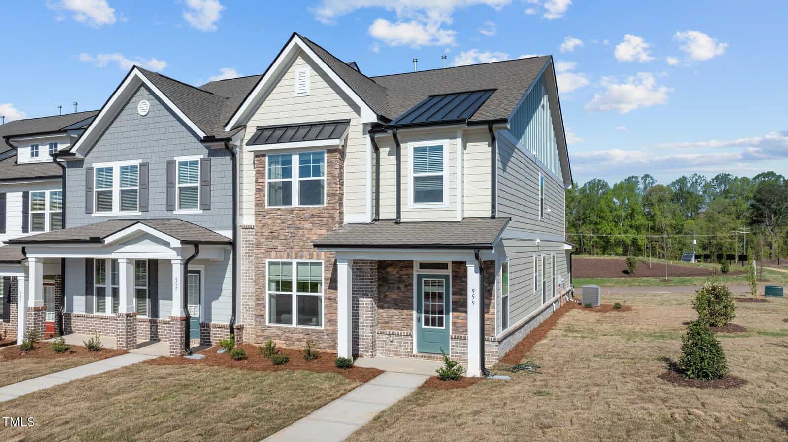 A two-story townhouse with gray and white siding, brick accents, a small covered front porch, and minimal landscaping. The yard has patchy grass, and there are trees and a clear sky in the background.