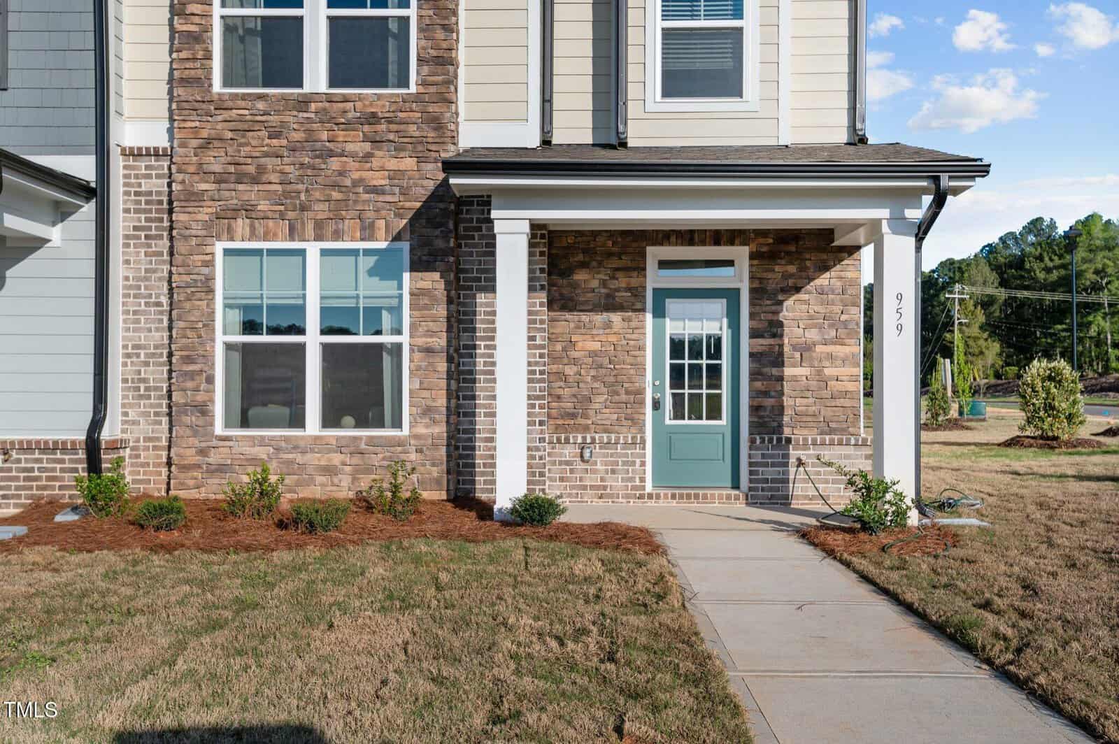 The image shows the front entrance of a two-story house with brick and siding exterior, a green front door with glass panels, several windows, a walkway, and landscaped shrubs in front.