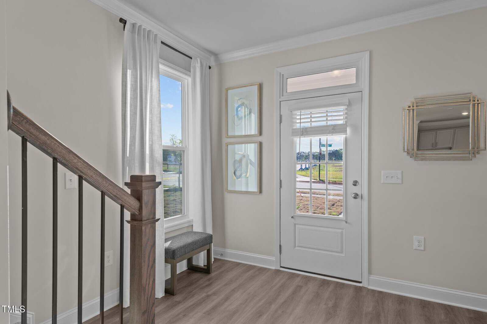 A small entryway with light wood flooring, a white front door with glass panels, a mirrored wall decor, two framed artworks, a window with white curtains, a gray bench, and a staircase with a wooden handrail.