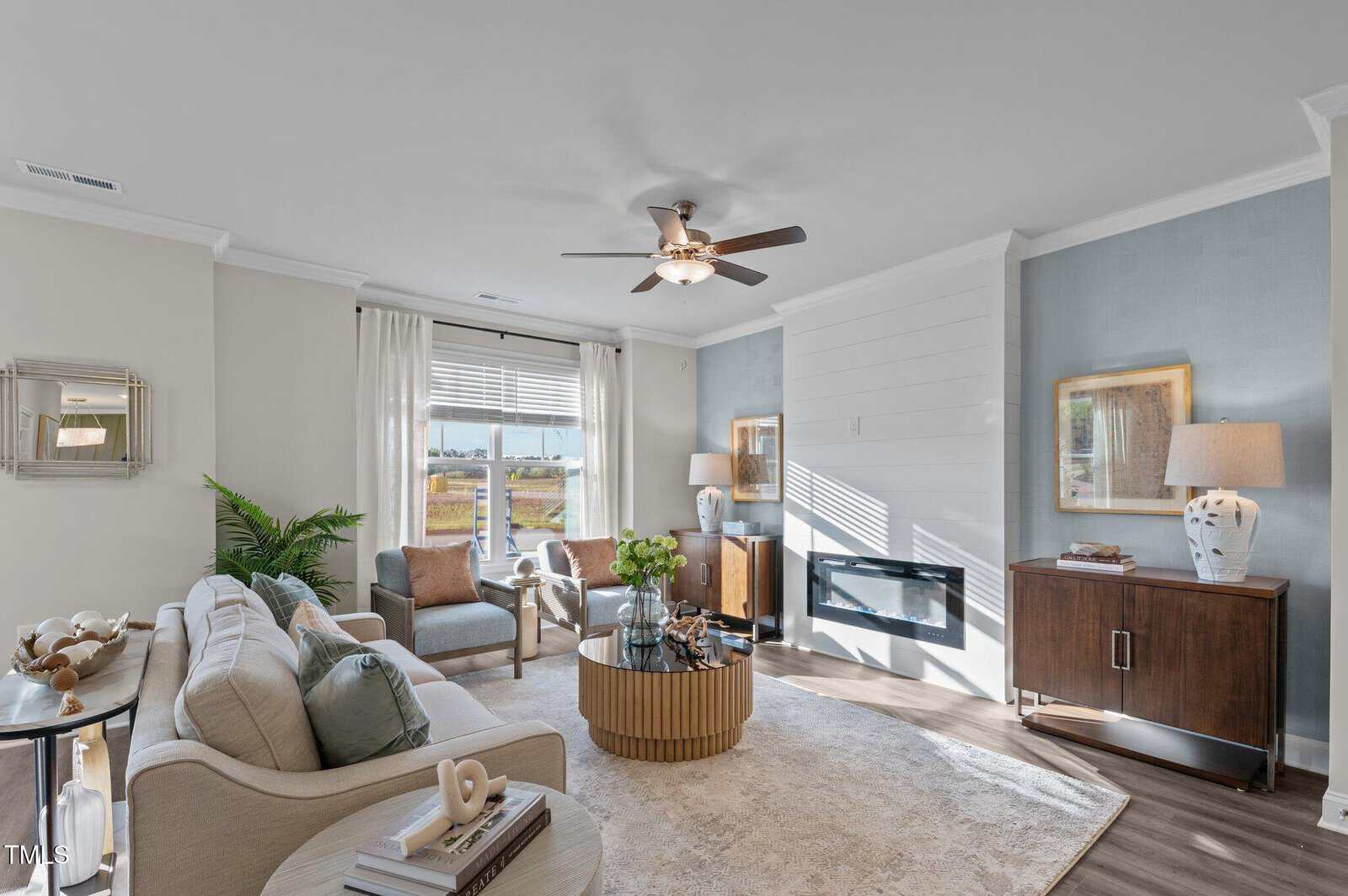 Bright, modern living room with neutral-toned furniture, large window, ceiling fan, fireplace, and decorative accents. Light blue accent wall, plants, and various decor create a cozy, inviting atmosphere.