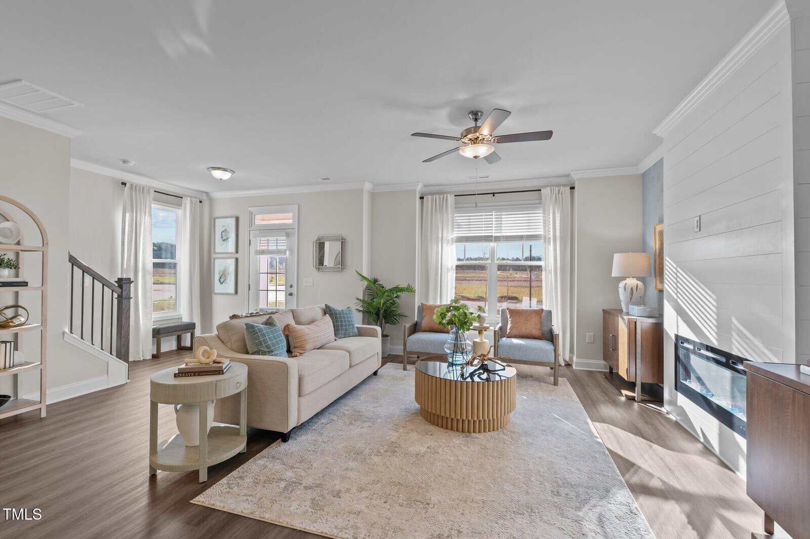 Bright living room with beige sofa, round coffee table, two accent chairs, large windows, ceiling fan, bookshelf, and electric fireplace. Neutral colors and modern decor create a welcoming, airy space with natural light.
